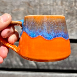 May include: A ceramic mug with a vibrant orange base, transitioning to a blue and brown rim. The handle is a contrasting brown. The mug is held in a hand, showcasing its colorful design. The mug is likely used for hot beverages.