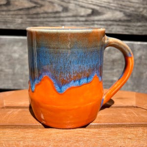May include: A ceramic mug featuring a glossy glaze with a vibrant orange base and a contrasting blue drip pattern cascading from the rim. The mug has a sturdy handle and a smooth, rounded body, showcasing a distinct colour transition where the blue glaze overlaps the bright orange surface.