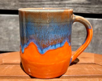 May include: A ceramic mug featuring a glossy glaze with a vibrant orange base and a contrasting blue drip pattern cascading from the rim. The mug has a sturdy handle and a smooth, rounded body, showcasing a distinct color transition where the blue glaze overlaps the bright orange surface.