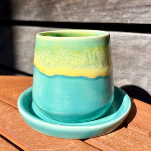 Op de afbeelding: Een keramieken beker en bijpassende schotel met een gladde, glanzende glazuurlaag. De beker heeft een levendige turquoise basis die bij de rand overgaat in een heldergeel en lichtgroen kleurverloop. De schotel heeft een effen turquoise kleur.