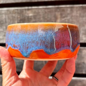 May include: A round ceramic bowl featuring a vibrant glaze with a bright orange base that transitions into a deep blue and purple drip pattern. The upper rim is finished in a warm golden-orange tone, creating a smooth, glossy surface texture.