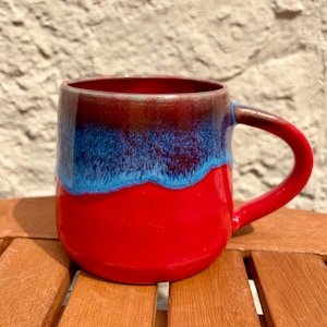 May include: A ceramic mug featuring a vibrant red base that transitions into a textured blue glaze near the rim, topped with a dark brown edge. The mug has a rounded body and a sturdy, curved handle extending from the right side of the vessel.