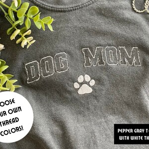 Puede incluir: Camiseta gris pimienta con bordado blanco que dice "DOG MOM" con una huella de pata debajo. El texto "CHOOSE YOUR OWN THREAD COLORS!" est&aacute; en un c&iacute;rculo blanco.