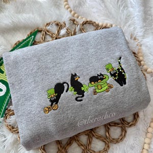 May include: A gray sweatshirt with an embroidered design of four black cats wearing green hats and holding shamrocks and gold coins. The text "@therachco" is embroidered on the bottom right corner of the sweatshirt.