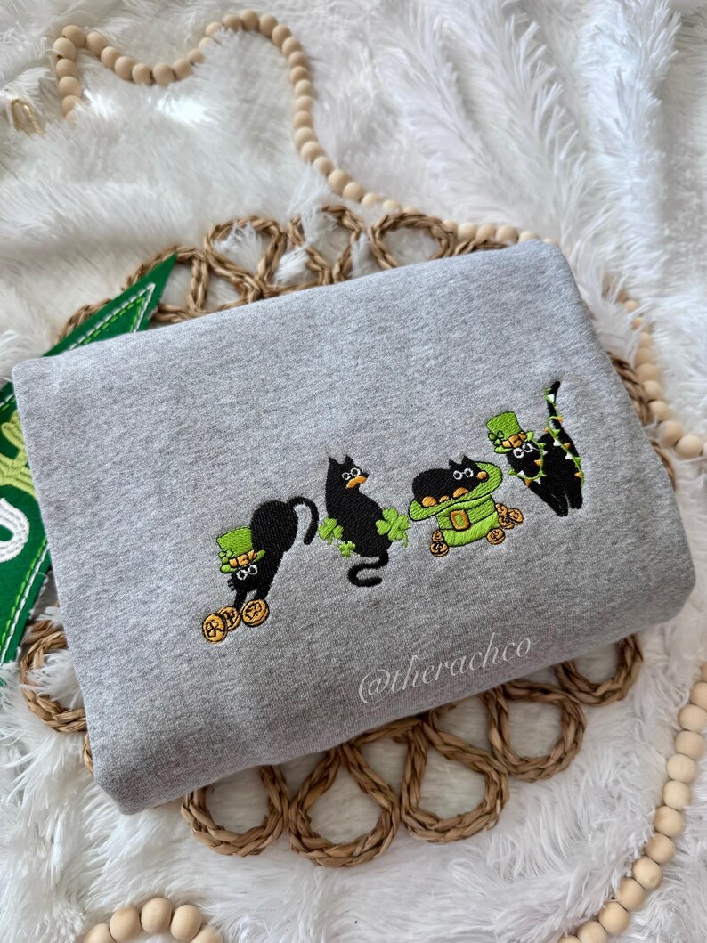May include: A gray sweatshirt with an embroidered design of four black cats wearing green hats and holding shamrocks and gold coins. The text "@therachco" is embroidered on the bottom right corner of the sweatshirt.