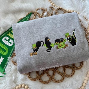 May include: A gray sweatshirt with an embroidered design of four black cats wearing green hats and holding gold coins. The cats are wearing shamrock necklaces. The text "@therachco" is embroidered on the bottom right corner of the sweatshirt.