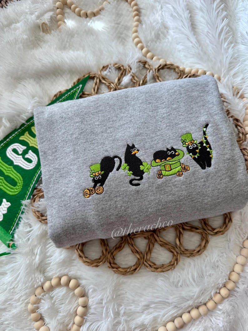 May include: A gray sweatshirt with an embroidered design of four black cats wearing green hats and holding gold coins. The cats are wearing shamrock necklaces. The text "@therachco" is embroidered on the bottom right corner of the sweatshirt.