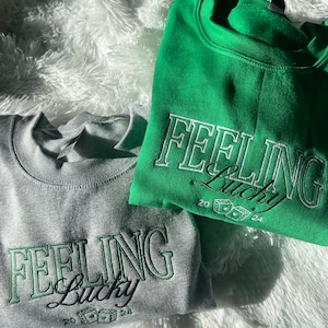 以下が含まれることがあります： 緑と白の刺繍で「Feeling Lucky」と書かれたスウェットシャツが2枚。スウェットシャツはグレーとグリーン。テキストは緑色のアウトラインで囲まれており、ドルマークのついたサイコロが含まれています。