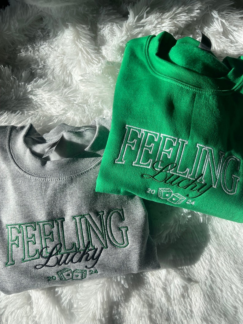以下が含まれることがあります： 緑と白の刺繍で「Feeling Lucky」と書かれたスウェットシャツが2枚。スウェットシャツはグレーとグリーン。テキストは緑色のアウトラインで囲まれており、ドルマークのついたサイコロが含まれています。