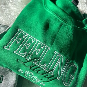 以下が含まれることがあります： 「Feeling Lucky」という白い刺繍の文字とサイコロ、20と24の数字が書かれた緑色のスウェットシャツです。