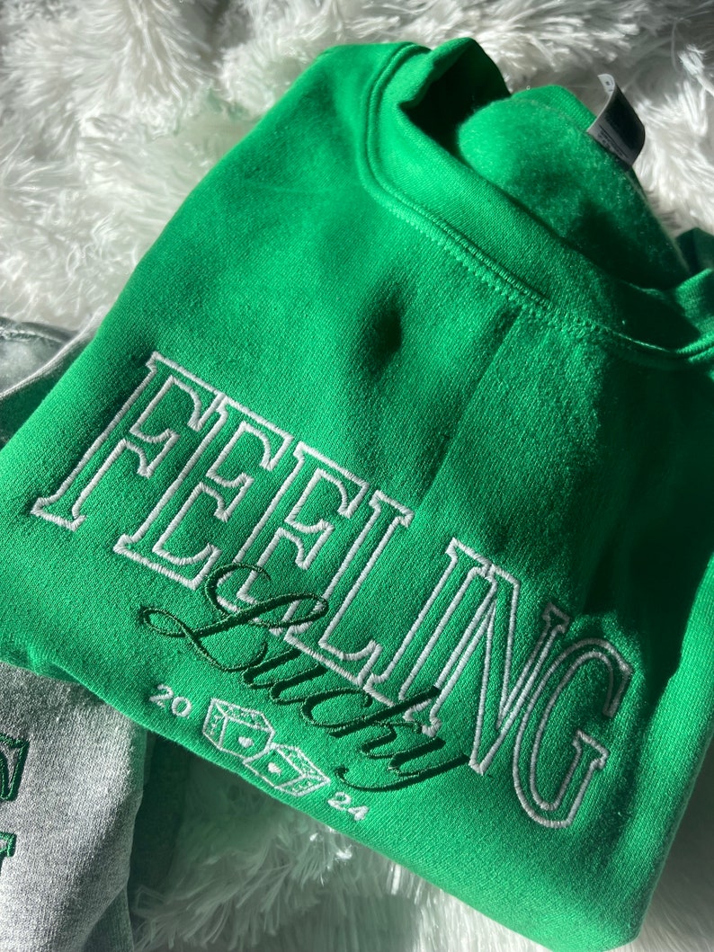 以下が含まれることがあります： 「Feeling Lucky」という白い刺繍の文字とサイコロ、20と24の数字が書かれた緑色のスウェットシャツです。