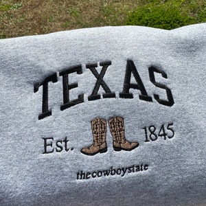 May include: Gray sweatshirt with black embroidered text that reads "TEXAS Est. 1845 the cowboystate" with a pair of brown cowboy boots.