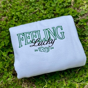 以下が含まれることがあります： 薄い青色のスウェットシャツに、「Feeling Lucky」という文字が緑色で刺繍されています。文字はレトロなフォントで、ハートのついたサイコロが2つ含まれています。スウェットシャツには、20と24の数字も刺繍されています。