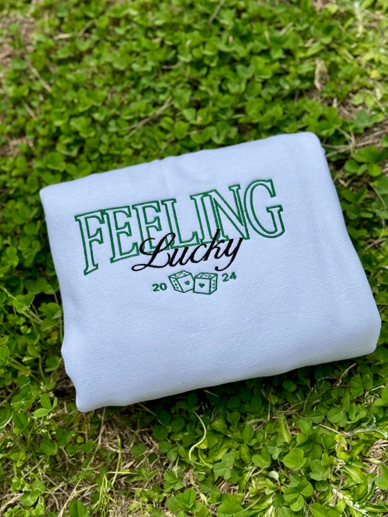 以下が含まれることがあります： 薄い青色のスウェットシャツに、「Feeling Lucky」という文字が緑色で刺繍されています。文字はレトロなフォントで、ハートのついたサイコロが2つ含まれています。スウェットシャツには、20と24の数字も刺繍されています。