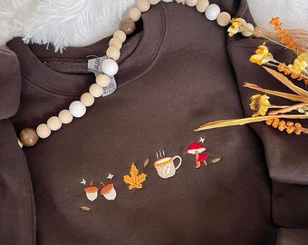 May include: A brown sweatshirt with an embroidered design of acorns, a leaf, a steaming mug, and a mushroom. The sweatshirt has a white and brown beaded necklace draped over it.