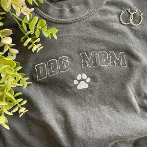 Puede incluir: Una camiseta gris oscura con las palabras "DOG MOM" bordadas en blanco con una huella de pata blanca debajo.