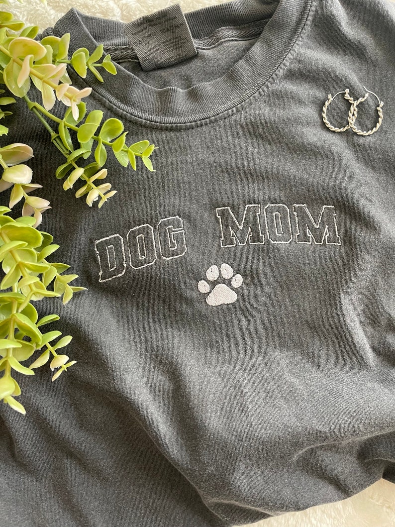 Puede incluir: Una camiseta gris oscura con las palabras "DOG MOM" bordadas en blanco con una huella de pata blanca debajo.
