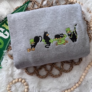 May include: A gray sweatshirt with an embroidered design of four black cats wearing green hats and holding gold coins. The cats are wearing green hats with shamrocks and are holding gold coins. The design is in a line across the front of the sweatshirt.