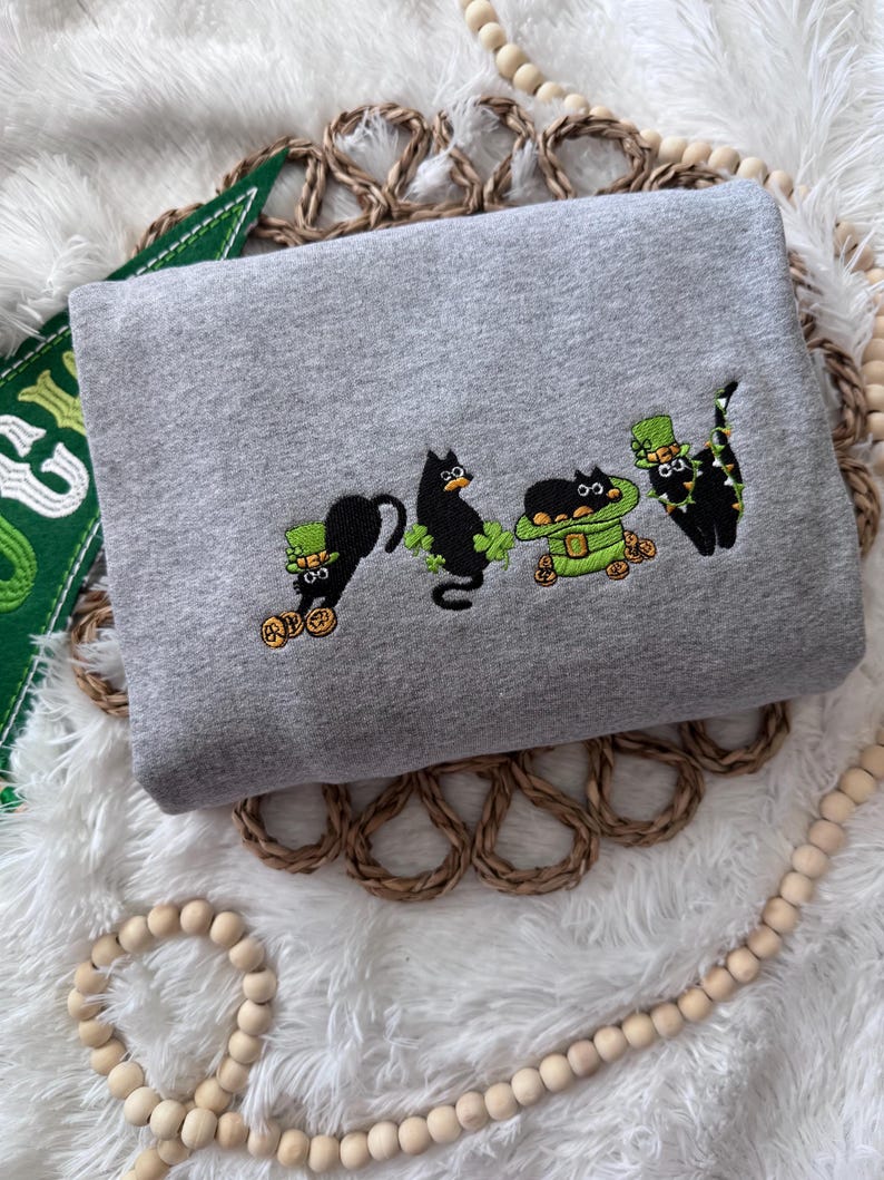 May include: A gray sweatshirt with an embroidered design of four black cats wearing green hats and holding gold coins. The cats are wearing green hats with shamrocks and are holding gold coins. The design is in a line across the front of the sweatshirt.