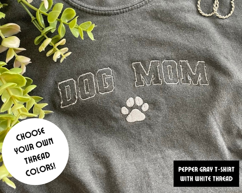 Puede incluir: Camiseta gris pimienta con bordado blanco que dice "DOG MOM" con una huella de pata debajo. El texto "CHOOSE YOUR OWN THREAD COLORS!" est&aacute; en un c&iacute;rculo blanco.