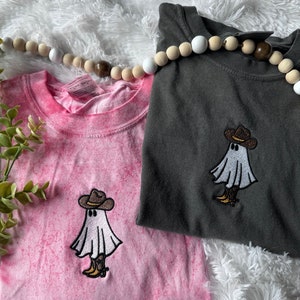 May include: Two t-shirts with a tie-dye pink and a dark gray color. Both shirts feature an embroidered design of a white ghost wearing a brown cowboy hat and boots.