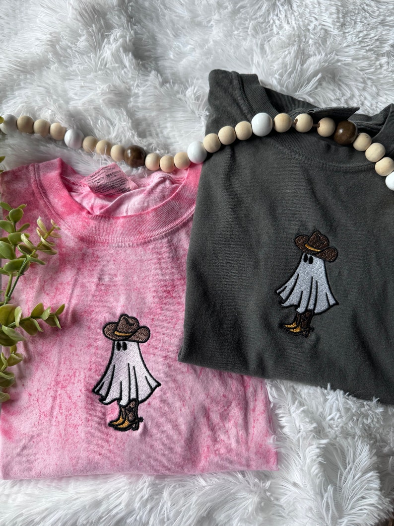 May include: Two t-shirts with a tie-dye pink and a dark gray color. Both shirts feature an embroidered design of a white ghost wearing a brown cowboy hat and boots.