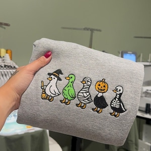May include: A heather gray sweatshirt featuring an embroidered design of five ducks in a row. The ducks are dressed in Halloween costumes: a witch with a broom, a green monster, a mummy, a pumpkin-headed duck, and a skeleton duck.