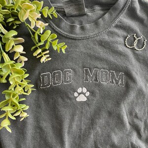 Puede incluir: Una camiseta gris con el texto "DOG MOM" bordado en blanco con una huella de pata blanca debajo del texto.