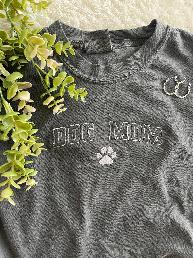 Puede incluir: Una camiseta gris con el texto "DOG MOM" bordado en blanco con una huella de pata blanca debajo del texto.