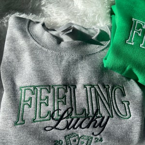 以下が含まれることがあります： グレーのトレーナーに、緑色の刺繍で「Feeling Lucky」という文字と、サイコロと数字の20と24が刺繍されています。