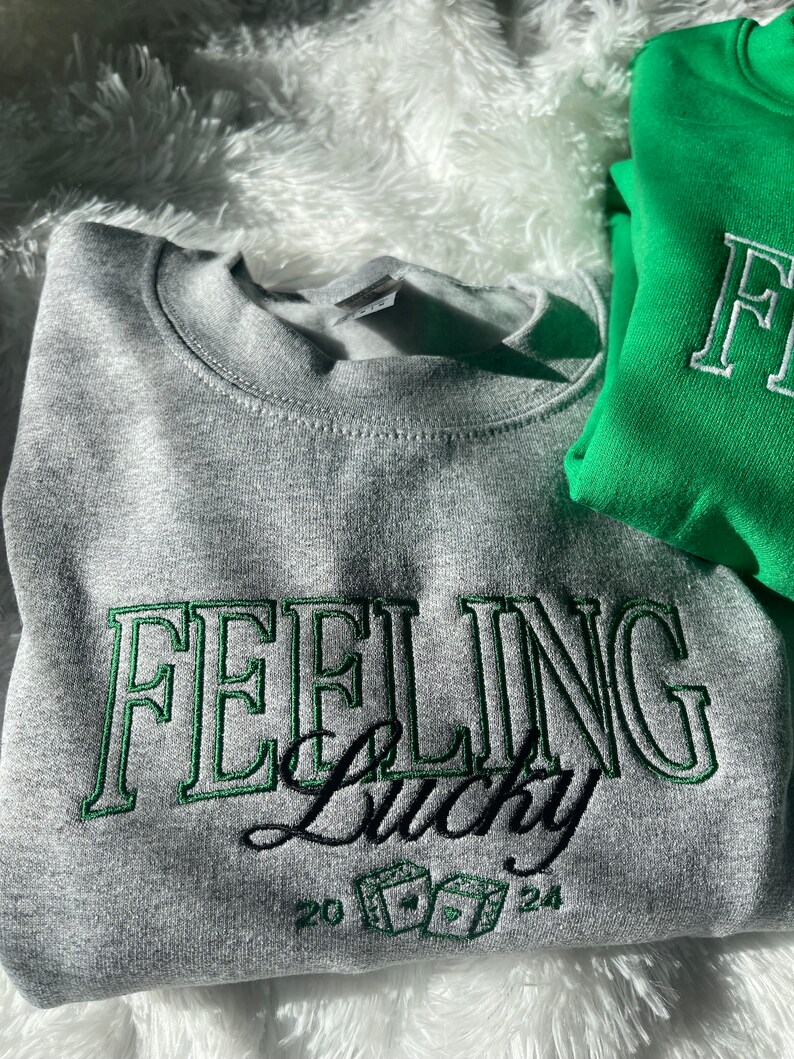 以下が含まれることがあります： グレーのトレーナーに、緑色の刺繍で「Feeling Lucky」という文字と、サイコロと数字の20と24が刺繍されています。