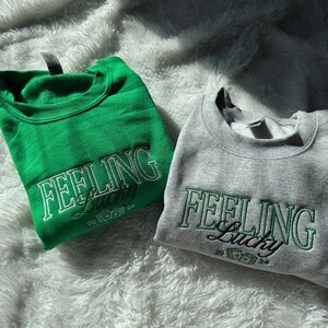 以下が含まれることがあります： 「Feeling Lucky」という文字とサイコロのグラフィックがプリントされたスウェットシャツが2枚。1枚は緑色、もう1枚は灰色です。
