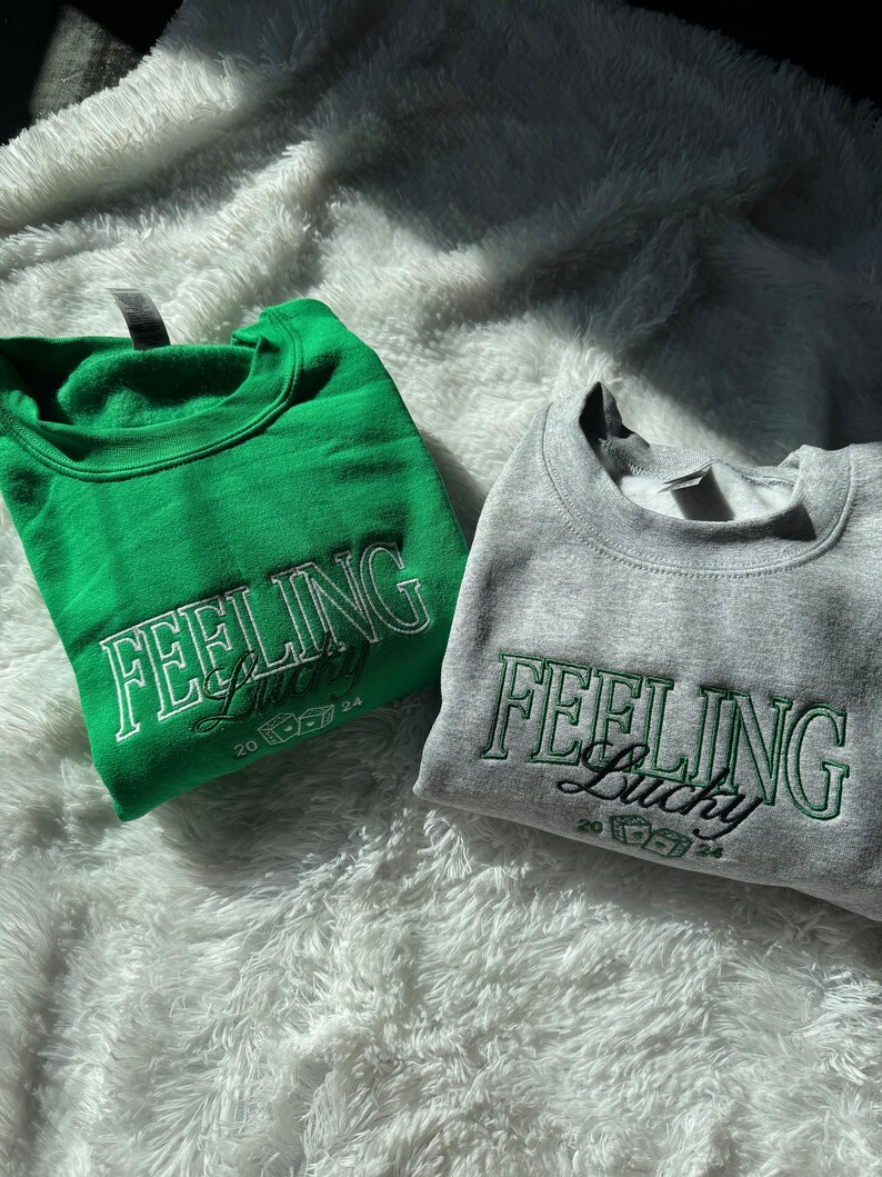 以下が含まれることがあります： 「Feeling Lucky」という文字とサイコロのグラフィックがプリントされたスウェットシャツが2枚。1枚は緑色、もう1枚は灰色です。