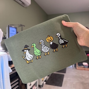 Puede incluir: Una sudadera verde oliva con un diseño bordado de cinco patos en fila. Los patos llevan disfraces de Halloween: una bruja con escoba, un pato verde, una momia, un pato con cabeza de calabaza y un pato con estampado de esqueleto.