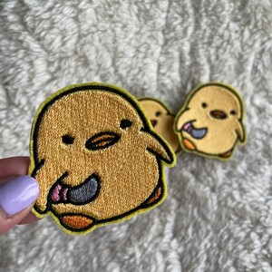 May include: Two yellow embroidered iron-on patches of a cartoon chick with a pink tongue sticking out. The chick on the left is holding a knife.