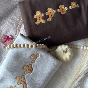 Może przedstawiać: Dwie bluzy z haftem w kształcie piernikowego ludzika. Górna bluza jest brązowa z czterema piernikowymi ludzikami, dolna bluza jest szara z trzema piernikowymi ludzikami.