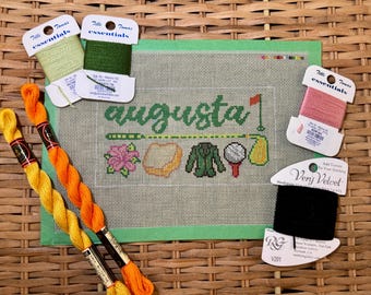 May include: Needlepoint canvas with the word "Augusta" in green script, featuring golf-themed icons. Colorful embroidery floss in shades of green, orange, yellow, pink, and black are arranged around the canvas.