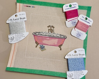May include: Needlepoint canvas with a pink bathtub design, surrounded by spools of thread in various colors. The canvas has a green border. The spools include pink, blue, and silver threads. The image is a close-up shot.