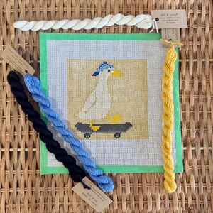 May include: A needlepoint canvas featuring a duck on a skateboard, with blue and yellow accents. The canvas is surrounded by green fabric and accompanied by skeins of blue, black, yellow, and white embroidery floss.