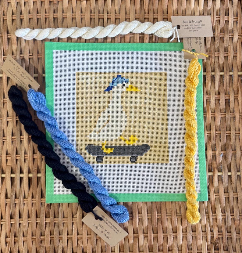 May include: A needlepoint canvas featuring a duck on a skateboard, with blue and yellow accents. The canvas is surrounded by green fabric and accompanied by skeins of blue, black, yellow, and white embroidery floss.