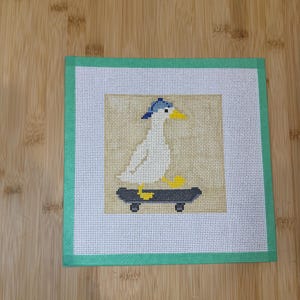 May include: Needlepoint art featuring a white seagull wearing a blue cap, riding a skateboard. The seagull has a yellow beak and feet. The artwork is set against a tan background, framed by a green border.