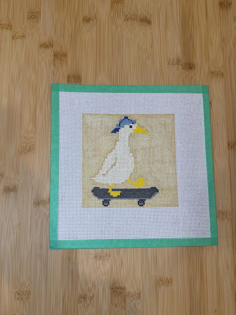 May include: Needlepoint art featuring a white seagull wearing a blue cap, riding a skateboard. The seagull has a yellow beak and feet. The artwork is set against a tan background, framed by a green border.
