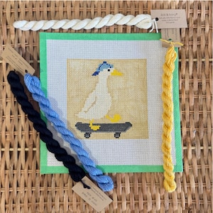 May include: Needlepoint canvas featuring a goose on a skateboard, with the text "A Silly Goose on the Scoot." The canvas is surrounded by green fabric and includes skeins of black, blue, and yellow embroidery floss. The background is a woven texture.