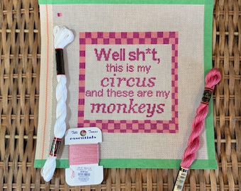 May include: A square needlepoint canvas featuring a pink and red checkered border and the text "Well sh*t, this is my circus and these are my monkeys" in dark pink. Included are two skeins of embroidery floss in white and pink, and a spool of pink thread.