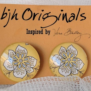 May include: A pair of yellow and white floral glass earrings with a blue center. The earrings are on a card that says "bjh Originals Inspired by Vera Bradley".