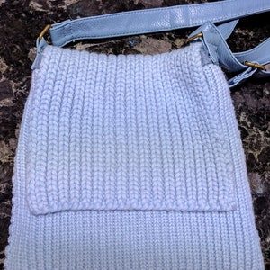 May include: A light blue knitted crossbody bag with a leather strap. The bag has a flap closure and a zippered pocket on the front.