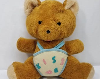 May include: A vintage brown teddy bear plush toy with a light blue diaper cover. The diaper has the word "KISS" and other shapes in pink and teal. The bear has a black nose and eyes, and tan ears.