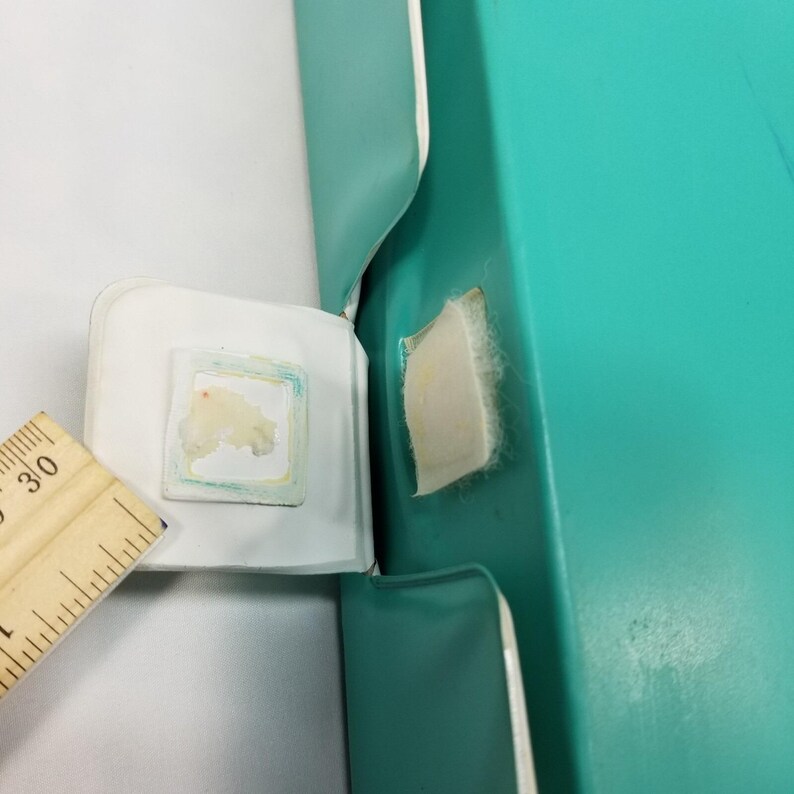 Op de afbeelding: Een close-up van een turquoise doos met een wit interieur. Een houten liniaal met het nummer 30 is gedeeltelijk zichtbaar. Een kleine, vierkante sticker met een kleurrijke rand is aan de doos bevestigd.