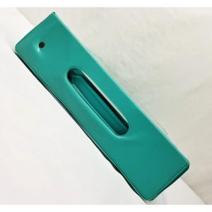 Op de afbeelding: Een turquoise rechthoekig object met een lange, ovale opening in het midden. Het object heeft een glanzende afwerking en een kleine, donkerrode stip op het bovenoppervlak. De opening onthult een geribbeld interieur.