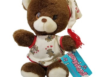 May include: A brown plush teddy bear wearing a cream-colored nightcap with a red tassel and a matching shirt featuring a repeating pattern of bears and red hearts. A blue rectangular tag attached to the bear displays the text Musicals by Applause.